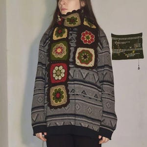 May include: A handmade crochet scarf featuring square floral motifs in shades of brown, red, green, and cream. The scarf is worn over a gray patterned sweater and black pants. The scarf's design includes a scalloped edge.