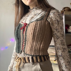 Peut inclure: Un gilet crocheté fait main avec un motif en tricot torsadé. Le gilet présente un panneau vert foncé à gauche, un panneau beige à droite et une fermeture à nouer rouge. L'ourlet inférieur est à rayures marron et beige. Le gilet est porté sur un chemisier à imprimé floral.