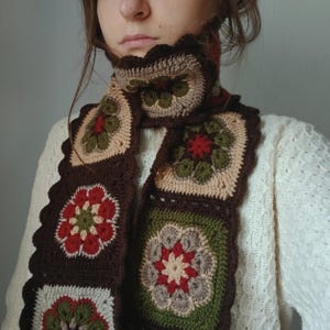 May include: A handmade crochet scarf featuring a floral pattern. The scarf is made of brown, beige, green, and red yarn. The scarf has a scalloped edge and is worn around the neck.