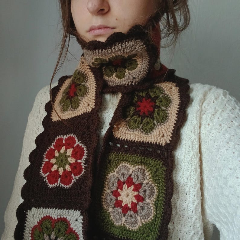 May include: A handmade crochet scarf featuring a floral pattern. The scarf is made of brown, beige, green, and red yarn. The scarf has a scalloped edge and is worn around the neck.