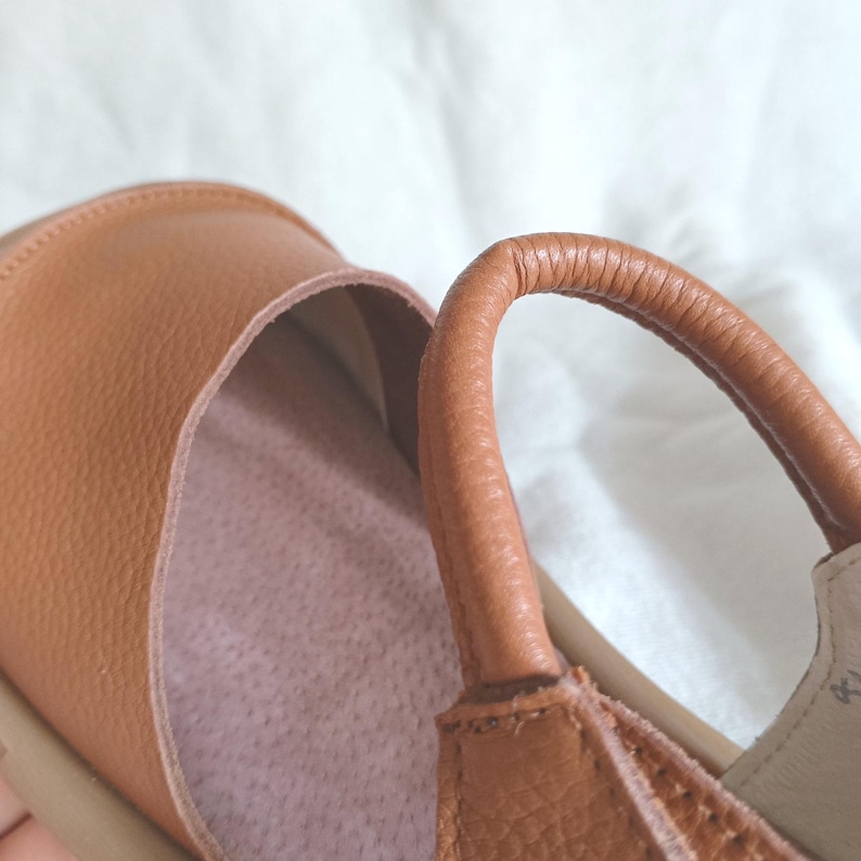 May include: A tan leather shoe featuring a textured grain finish, a rounded toe area, and a curved ankle strap. The interior of the shoe is lined with a light pink, soft-textured material, and the outer construction shows visible stitching details.