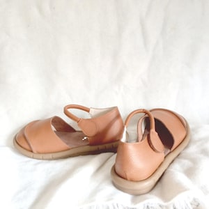 May include: A pair of tan leather sandals with a textured finish, featuring a rounded toe, a secure ankle strap with a side closure, and a flat, flexible rubber sole. The design includes a wide front strap and a supportive heel counter.