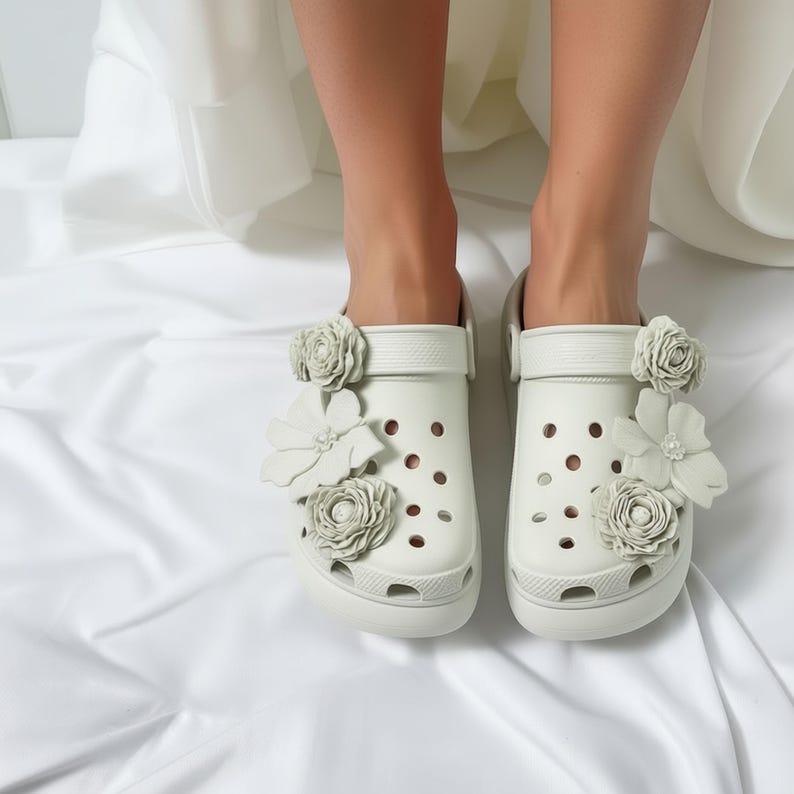 “Close-up of a pair of white slip-on clogs decorated with large white floral embellishments, worn by a person standing on white fabric.”