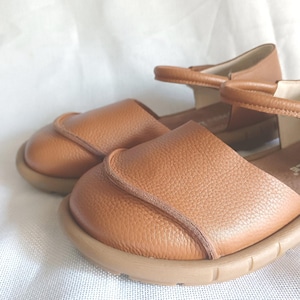 May include: A pair of tan leather sandals featuring a rounded closed-toe design with a textured grain finish. The footwear includes a thin ankle strap with a buckle closure and a flexible, flat rubber sole in a matching tan color.