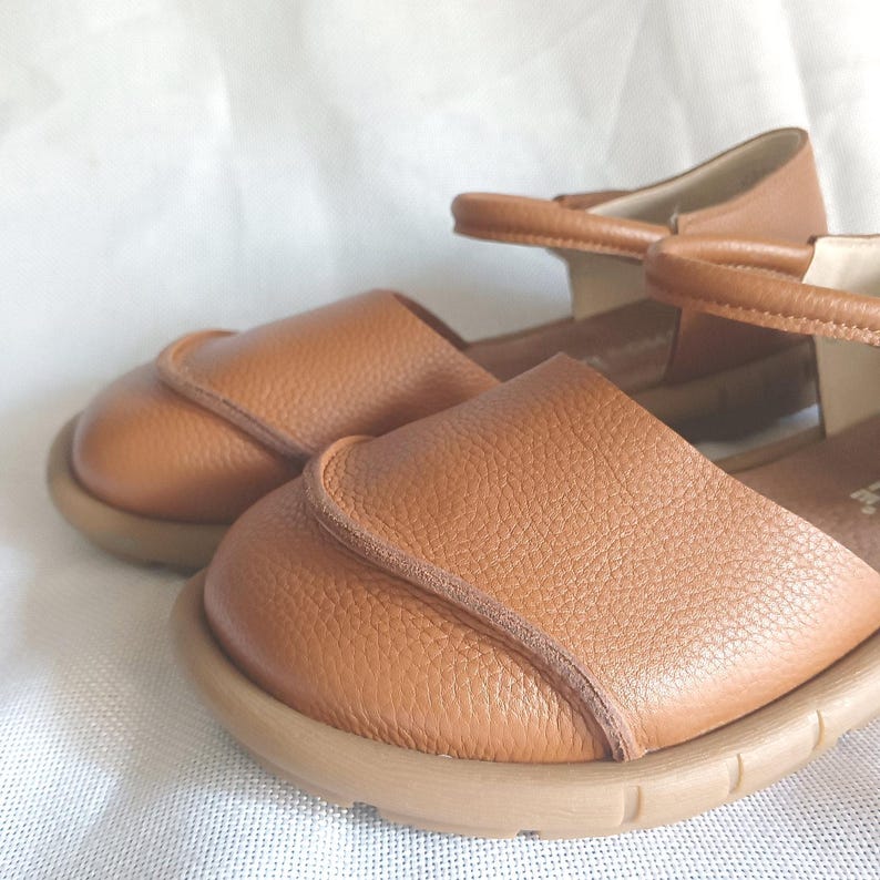 May include: A pair of tan leather sandals featuring a rounded closed-toe design with a textured grain finish. The footwear includes a thin ankle strap with a buckle closure and a flexible, flat rubber sole in a matching tan color.