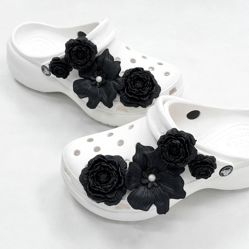 Pu&ograve; includere: Un paio di zoccoli bianchi adornati con grandi abbellimenti floreali neri. I fiori includono rose e altri fiori stilizzati, alcuni con un centro perlato. Gli zoccoli sono su uno sfondo bianco.