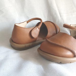 May include: A pair of tan leather shoes with a pebbled texture, featuring a rounded toe, a low-profile design, and a single strap across the instep with a hook-and-loop closure. The shoes have a flexible, flat, light brown rubber sole.