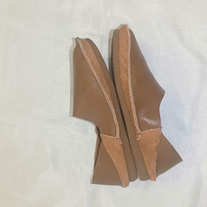 May include: A pair of brown leather slippers. The slippers have a rounded toe and a low heel. The upper is made of a textured brown leather, and the sole is a lighter brown. The heel and toe are reinforced with a suede-like material.