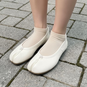May include: A pair of white split-toe ballet flats with a small bow detail on the vamp. The shoes are worn with beige ankle socks. The flats have a rounded toe and a low heel, and are made of a smooth, leather-like material.