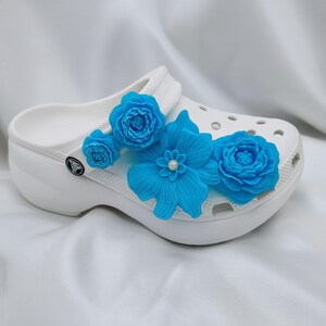 Pu&ograve; includere: Zoccoli bianchi adornati con vivaci decorazioni floreali turchesi. I fiori decorativi includono un grande fiore a pi&ugrave; petali e piccoli accenti a forma di rosa. Gli zoccoli hanno una suola platform e il classico logo Crocs.