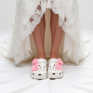 “White slip-on clogs decorated with pink flowers charms, worn by a person in a white lace dress standing on a white surface.”