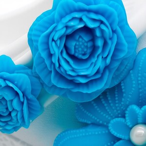 Pu&ograve; includere: Primo piano di decorazioni floreali blu vibranti. L'immagine mostra fiori scolpiti in dettaglio con petali a strati e un accento a forma di perla. I fiori sono ambientati su uno sfondo bianco, evidenziando il loro colore brillante e il design intricato.