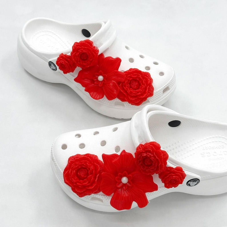 Pu&ograve; includere: Zoccoli bianchi adornati con vivaci decorazioni floreali rosse. Gli zoccoli presentano un design classico con molteplici fori di ventilazione e un cinturino al tallone girevole. I fiori rossi sono raggruppati sulla parte superiore degli zoccoli, aggiungendo un tocco di colore ed eleganza.