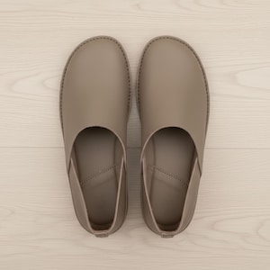May include: A pair of taupe-colored leather slip-on shoes. The shoes have a rounded toe and a low profile, with a stitched seam around the edge. The design is minimalist and the shoes appear to be unworn.
