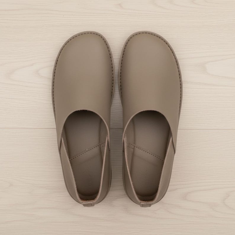 May include: A pair of taupe-colored leather slip-on shoes. The shoes have a rounded toe and a low profile, with a stitched seam around the edge. The design is minimalist and the shoes appear to be unworn.
