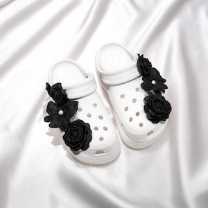 Pu&ograve; includere: Un paio di zoccoli bianchi adornati con decorazioni floreali nere. Gli zoccoli presentano un design classico con fori di ventilazione e una suola strutturata. I fiori neri aggiungono un tocco di eleganza alle calzature casual.