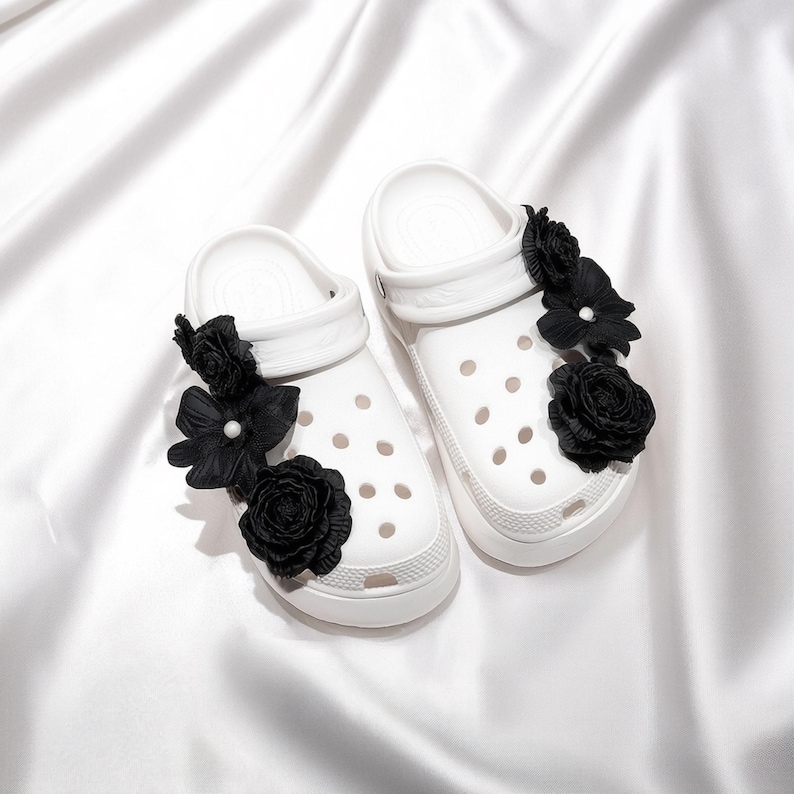 Pu&ograve; includere: Un paio di zoccoli bianchi adornati con decorazioni floreali nere. Gli zoccoli presentano un design classico con fori di ventilazione e una suola strutturata. I fiori neri aggiungono un tocco di eleganza alle calzature casual.