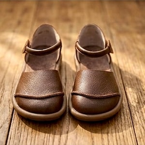 May include: A pair of brown leather shoes with a textured surface, featuring a rounded toe, a closed heel, and a thin ankle strap with a buckle closure. The shoes have a flat sole and a smooth interior lining, presented in a front-facing orientation.