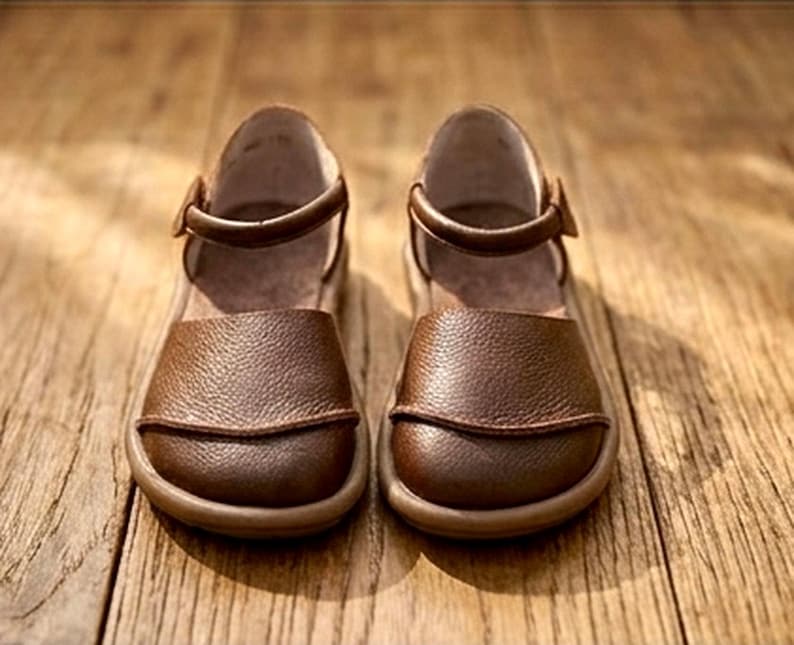 May include: A pair of brown leather shoes with a textured surface, featuring a rounded toe, a closed heel, and a thin ankle strap with a buckle closure. The shoes have a flat sole and a smooth interior lining, presented in a front-facing orientation.