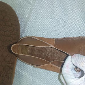 May include: Close-up of a brown leather shoe with a textured sole. The shoe's upper is made of soft, tanned leather, and the sole has a geometric pattern. The shoe is being held by a gloved hand.