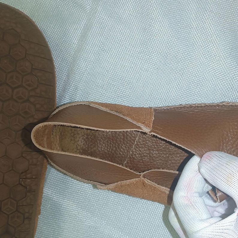 May include: Close-up of a brown leather shoe with a textured sole. The shoe's upper is made of soft, tanned leather, and the sole has a geometric pattern. The shoe is being held by a gloved hand.