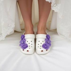“White slip-on clogs decorated with purple flowers charms, worn by a person in a white lace dress standing on a white surface.”