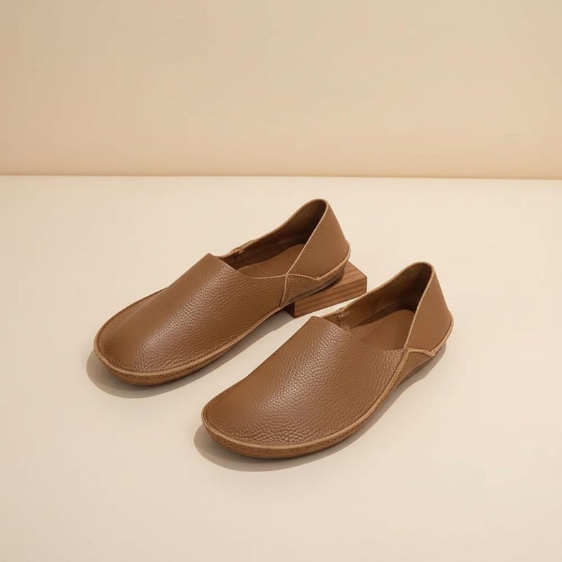May include: A pair of brown leather slip-on shoes. The shoes have a rounded toe and a low profile, with a textured leather upper and a light brown sole. The shoes are displayed on a light-colored surface.