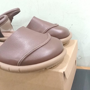 May include: A pair of taupe leather sandals with a rounded toe, a smooth upper panel, and a thick, textured beige rubber sole. The sandals feature a heel strap and are positioned on top of a plain, light brown cardboard shoe box.