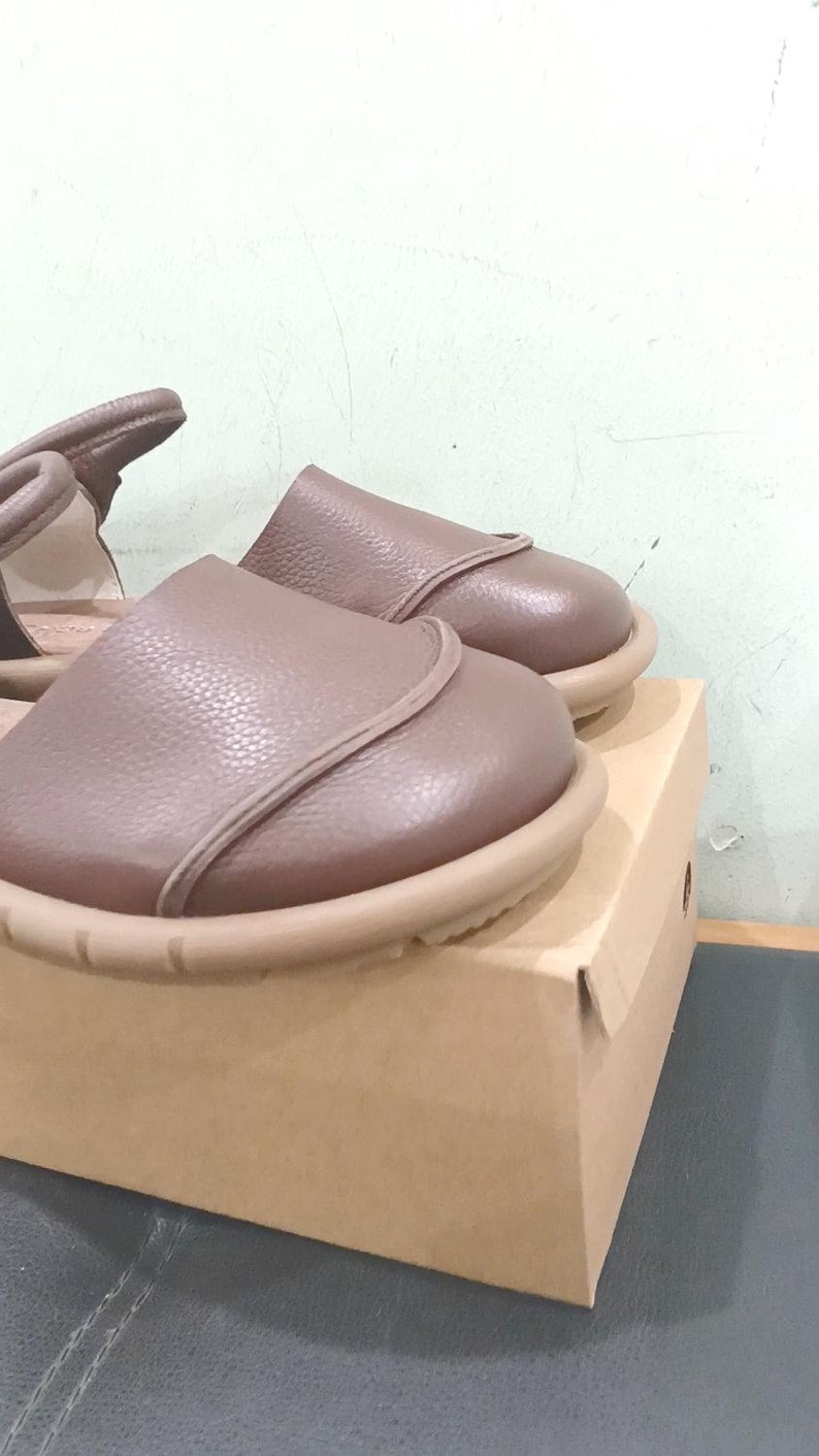 May include: A pair of taupe leather sandals with a rounded toe, a smooth upper panel, and a thick, textured beige rubber sole. The sandals feature a heel strap and are positioned on top of a plain, light brown cardboard shoe box.