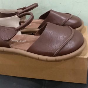 May include: A pair of brown leather sandals with a rounded toe, a textured surface, and a strap that secures around the ankle. The sandals feature a tan-colored sole and a light-colored interior lining with the text Hou Le visible on the insole.