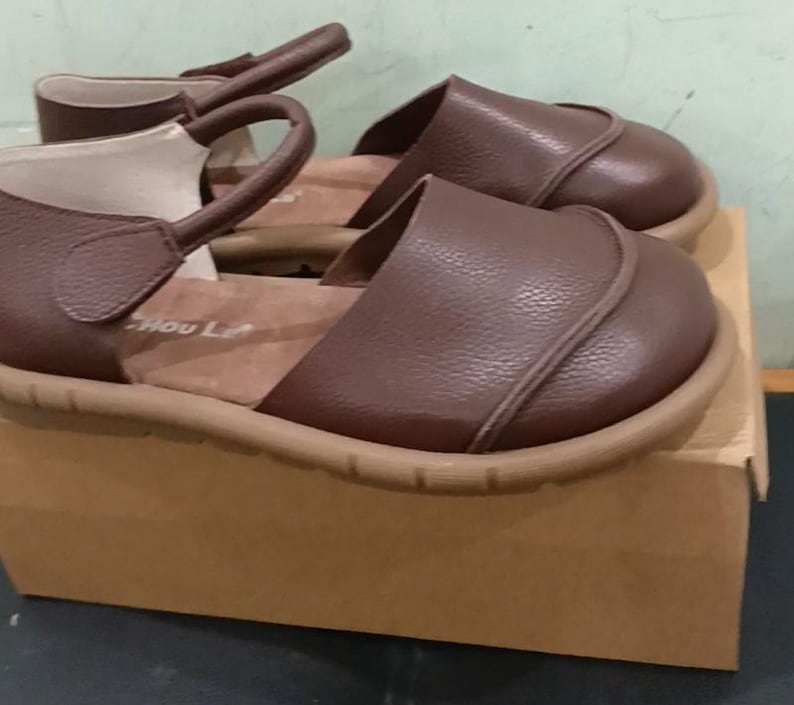 May include: A pair of brown leather sandals with a rounded toe, a textured surface, and a strap that secures around the ankle. The sandals feature a tan-colored sole and a light-colored interior lining with the text Hou Le visible on the insole.