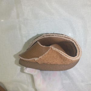 May include: A flexible, brown leather shoe, folded to show its construction. The shoe features a light brown suede-like interior and a darker brown sole. The image highlights the shoe's soft, pliable material and design.