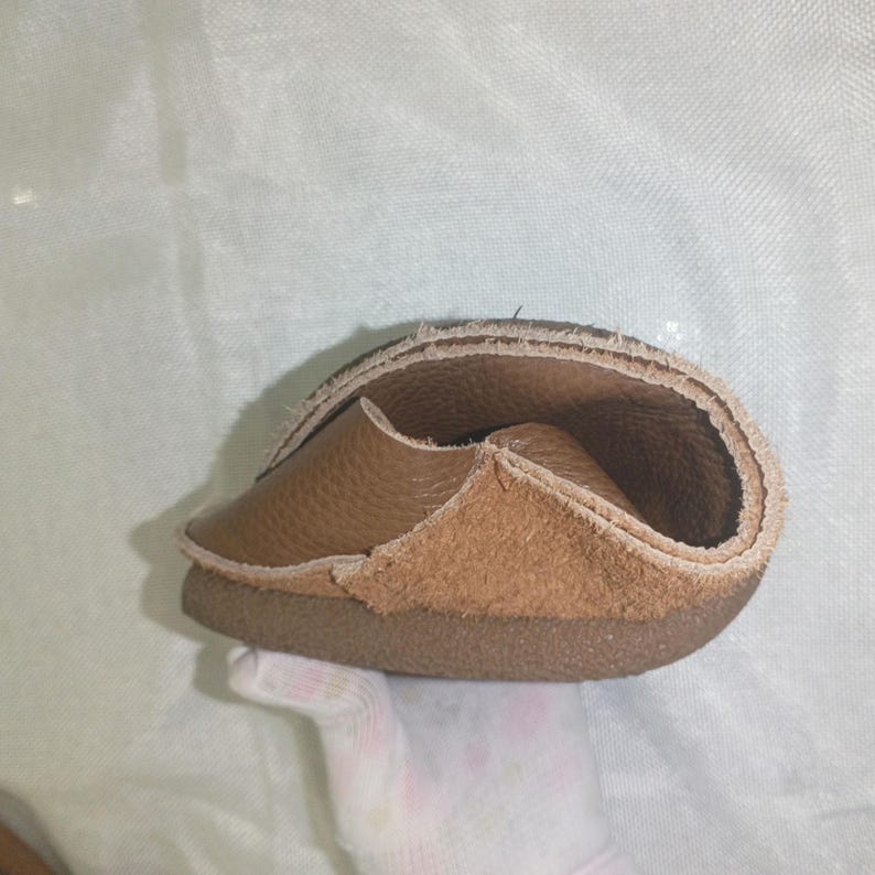 May include: A flexible, brown leather shoe, folded to show its construction. The shoe features a light brown suede-like interior and a darker brown sole. The image highlights the shoe's soft, pliable material and design.