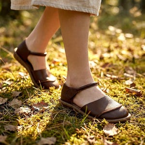 May include: A pair of dark brown leather sandals featuring a rounded toe, a textured upper panel, and a thin ankle strap with a side closure. The sandals have a flat, flexible sole and are worn with a light-colored fabric garment hem visible above.