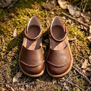 May include: A pair of brown leather shoes with a textured surface, featuring a rounded toe, a low-cut opening, and a single strap across the top with a side closure. The shoes have a tan-colored sole and a smooth interior lining.