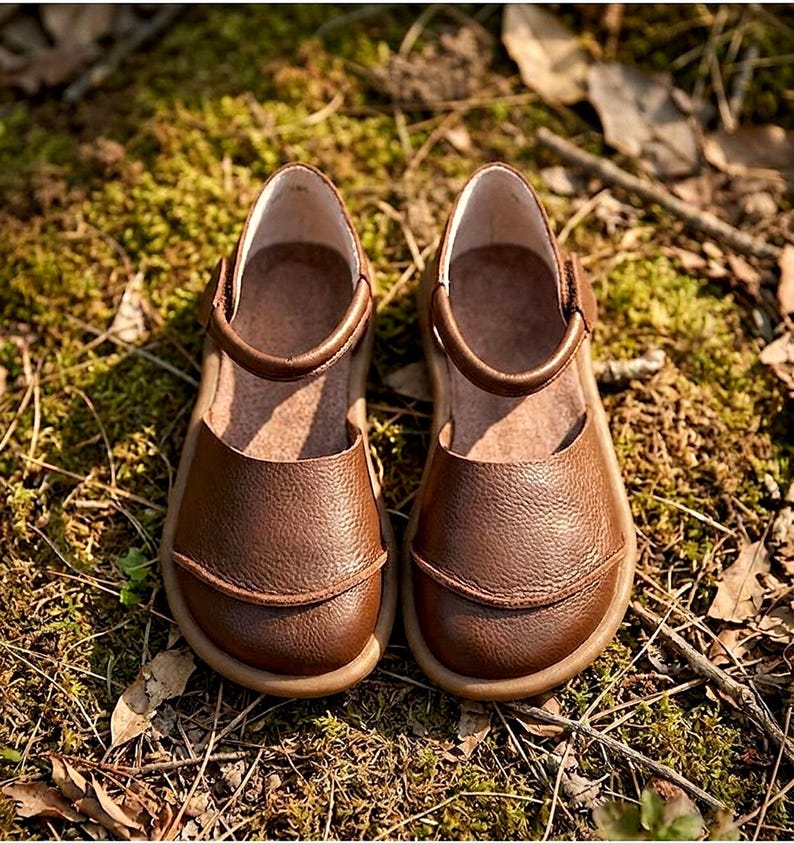 May include: A pair of brown leather shoes with a textured surface, featuring a rounded toe, a low-cut opening, and a single strap across the top with a side closure. The shoes have a tan-colored sole and a smooth interior lining.