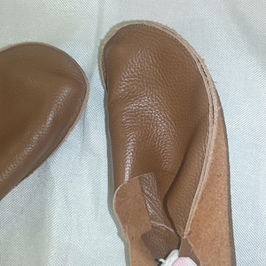 May include: A pair of brown leather baby shoes. The shoes have a soft, textured appearance and a simple design. The soles are a lighter shade of brown, and the shoes appear to be handmade. The shoes are designed for comfort and flexibility.