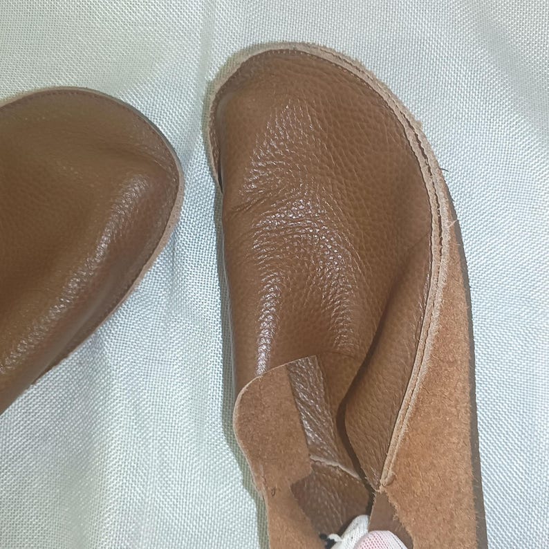 May include: A pair of brown leather baby shoes. The shoes have a soft, textured appearance and a simple design. The soles are a lighter shade of brown, and the shoes appear to be handmade. The shoes are designed for comfort and flexibility.