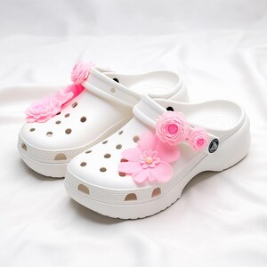 Pu&ograve; includere: Zoccoli bianchi con plateau decorati con fiori rosa. Gli zoccoli presentano un design classico con fori di ventilazione e cinturino al tallone girevole. I fiori rosa aggiungono un tocco giocoso alle calzature casual.