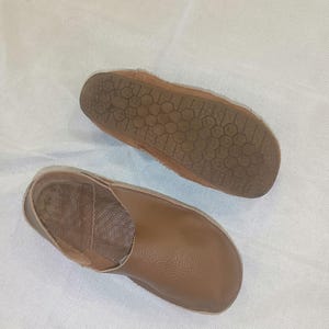 May include: A pair of brown leather slippers. One slipper is shown from the sole, revealing a textured pattern. The other slipper is shown from the top, displaying a smooth, rounded design. The slippers appear to be handmade.