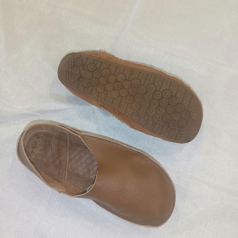 May include: A pair of brown leather slippers. One slipper is shown from the sole, revealing a textured pattern. The other slipper is shown from the top, displaying a smooth, rounded design. The slippers appear to be handmade.
