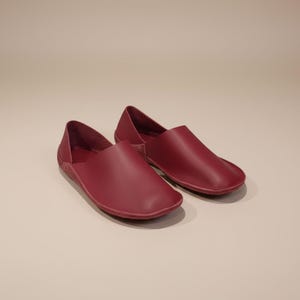 May include: A pair of burgundy leather slip-on shoes. The shoes have a rounded toe and a low profile, with a simple, minimalist design. The leather appears smooth and the color is a deep, rich red.