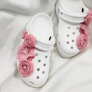 Pu&ograve; includere: Zoccoli bianchi adornati con decorazioni floreali rosa. Gli zoccoli presentano fori multipli e un cinturino. I fiori rosa sono raggruppati sul lato di ogni zoccolo, aggiungendo un tocco decorativo alle calzature.