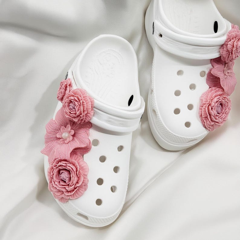 Pu&ograve; includere: Zoccoli bianchi adornati con decorazioni floreali rosa. Gli zoccoli presentano fori multipli e un cinturino. I fiori rosa sono raggruppati sul lato di ogni zoccolo, aggiungendo un tocco decorativo alle calzature.