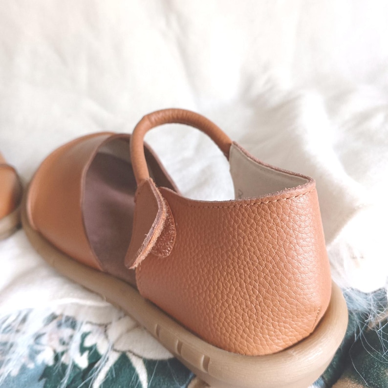 May include: A tan leather shoe with a textured pebble grain finish, featuring a rounded toe, a single strap across the instep with a side closure, and a thick, light-colored rubber sole. The interior lining is a smooth, light-colored material.
