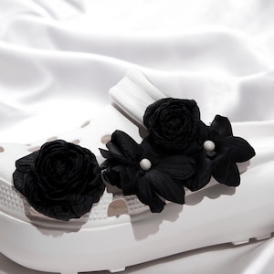 Pu&ograve; includere: Zoccoli bianchi adornati con decorazioni floreali nere. I fiori includono una rosa e diversi fiori a pi&ugrave; petali, alcuni con centri di perle bianche. Gli zoccoli poggiano su una superficie bianca e setosa.