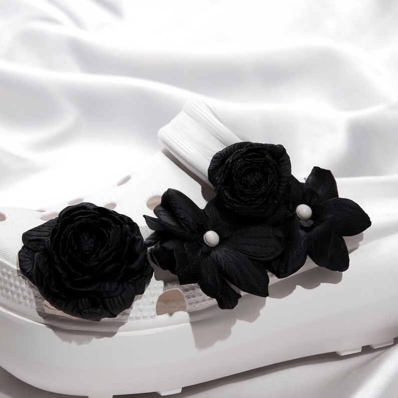 Pu&ograve; includere: Zoccoli bianchi adornati con decorazioni floreali nere. I fiori includono una rosa e diversi fiori a pi&ugrave; petali, alcuni con centri di perle bianche. Gli zoccoli poggiano su una superficie bianca e setosa.