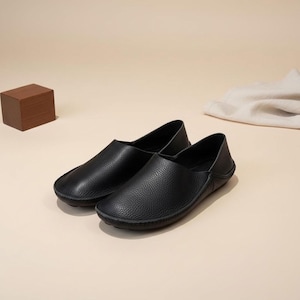 May include: A pair of black leather slip-on shoes with a textured surface. The shoes have a rounded toe and a low profile, suitable for casual wear. The background is a neutral beige.