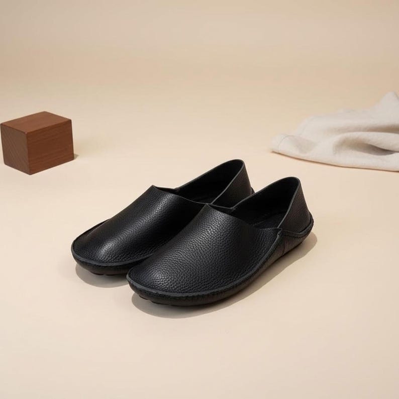 May include: A pair of black leather slip-on shoes with a textured surface. The shoes have a rounded toe and a low profile, suitable for casual wear. The background is a neutral beige.