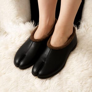 Könnte beinhalten: Schwarze Leder-Zehenslipper mit braunem Kunstfellfutter. Die Slipper haben ein einzigartiges Design mit einem separaten Fach für den großen Zeh. Die Schuhe sind auf Komfort und Wärme ausgelegt.